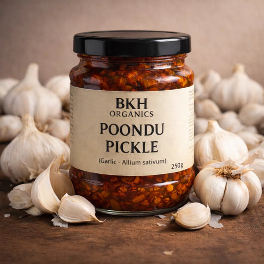 Jar of BKH Organics Poondu Pickle surrounded by garlic bulbs on a wooden surface.