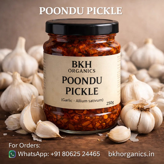 Jar of BKH Organics Poondu Pickle surrounded by garlic cloves on a wooden surface.