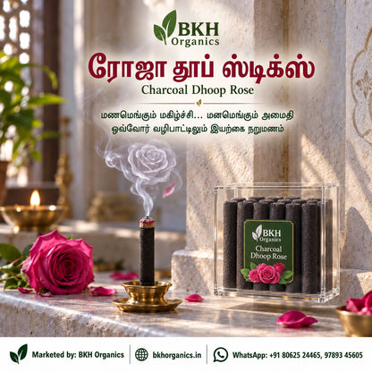 BKH Organics Charcoal Dhoop Rose advertisement showing a burning incense stick in a brass holder and a clear box of dark charcoal sticks with a rose label, set on a stone surface with a rose, petals, and lit diya in a real interior setting. BKH Organics Charcoal Dhoop Rose Sticks (தமிழ்: ரோஜா தூப் ஸ்டிக்ஸ், తెలుగు: గులాబీ ధూప్ స్టిక్స్, ಕನ್ನಡ: ಗುಲಾಬಿ ಧೂಪ್ ಕಡ್ಡಿಗಳು, മലയാളം: റോസ് ധൂപം / ധൂപ് സ്റ്റിക്കുകൾ, ತುಳು: ರೋಸ್ ಧೂಪ್ದ ಕಡ್ಡಿಕುಲು) lit on a brass holder with fragrant rose smoke and acrylic container box